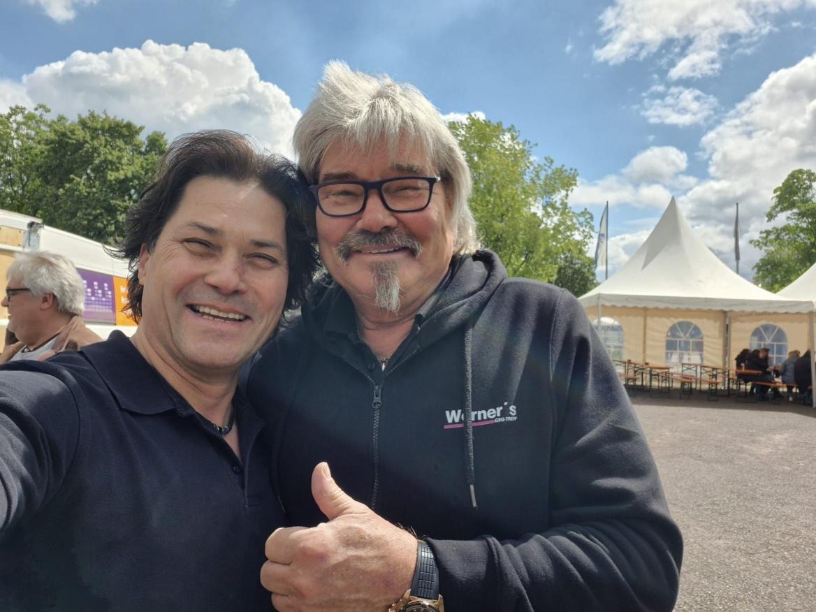 Zwei Männer machen ein Selfie im Freien, einer lächelt und zeigt den Daumen hoch.