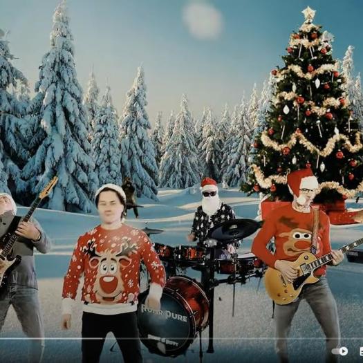 Eine Band spielt vor einer verschneiten Waldkulisse, die weihnachtliche Dekorationen wie einen großen Weihnachtsbaum und ein Lebkuchenhaus beinhaltet.